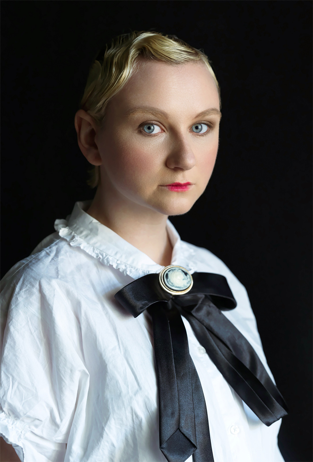 Milena Kaltenbach in weißer Bluse mit schwarzer Schleife und Kamee vor dunklem Grund, Fashion-Strecke