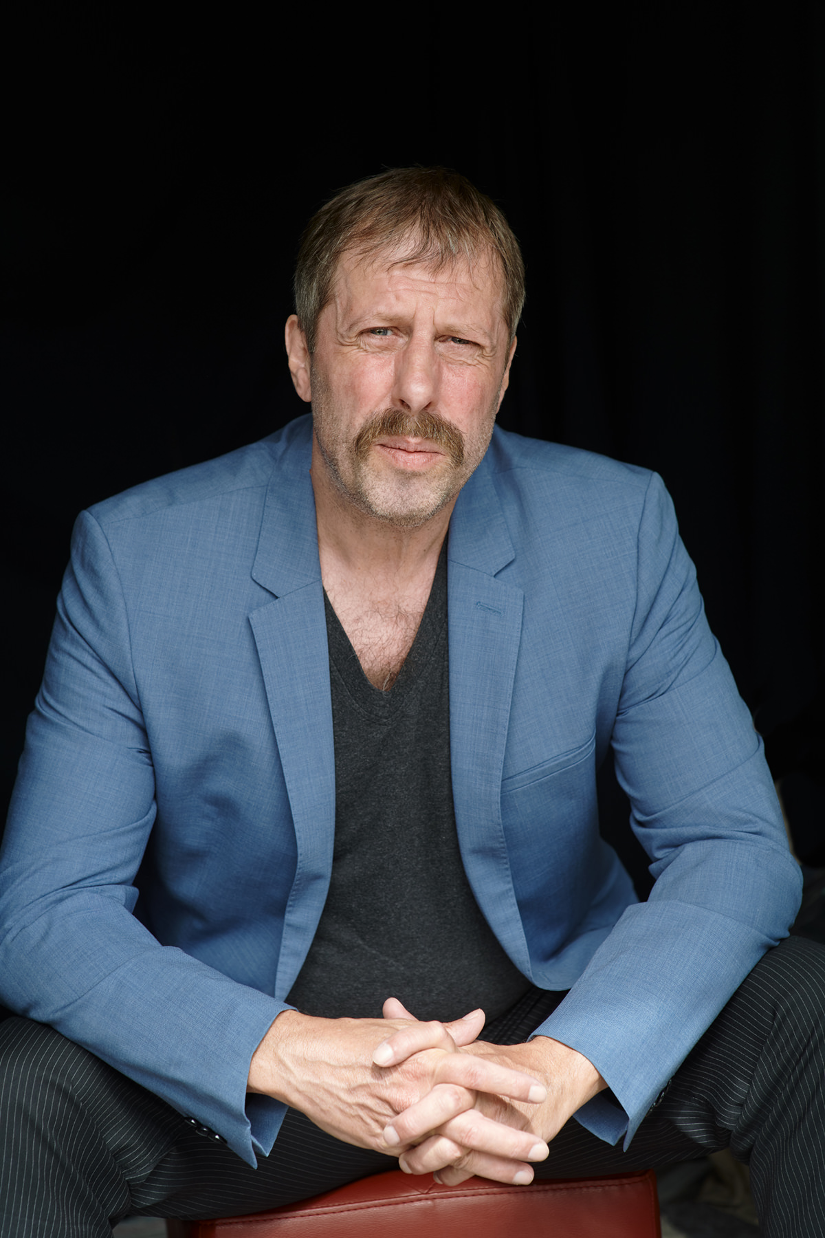 Oliver Korittke im hellblauen Sakko vor schwarzem Vorhang, sitzend mit gefalteten Händen