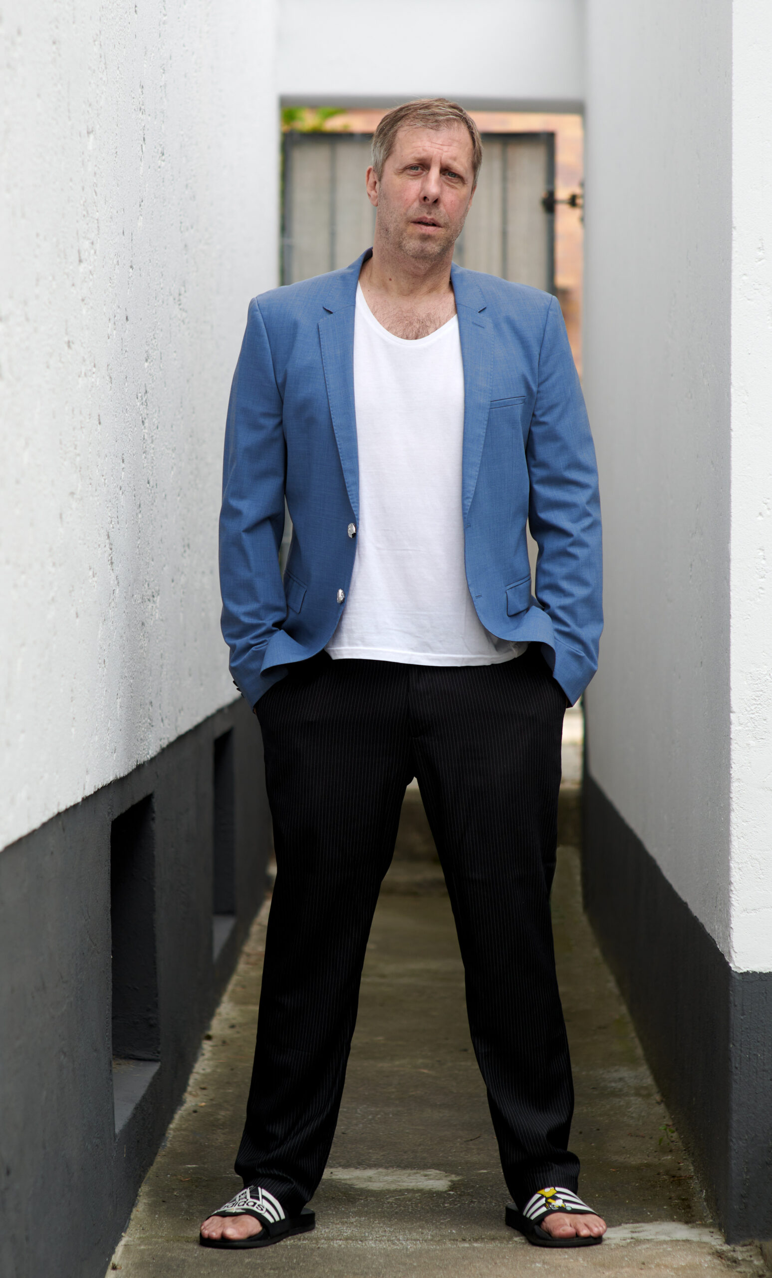 Oliver Korittke im hellblauen Sakko und weißen T-Shirt in einem schmalen weißen Durchgang, Hände in den Hosentaschen, Porträtfotografie Berlin